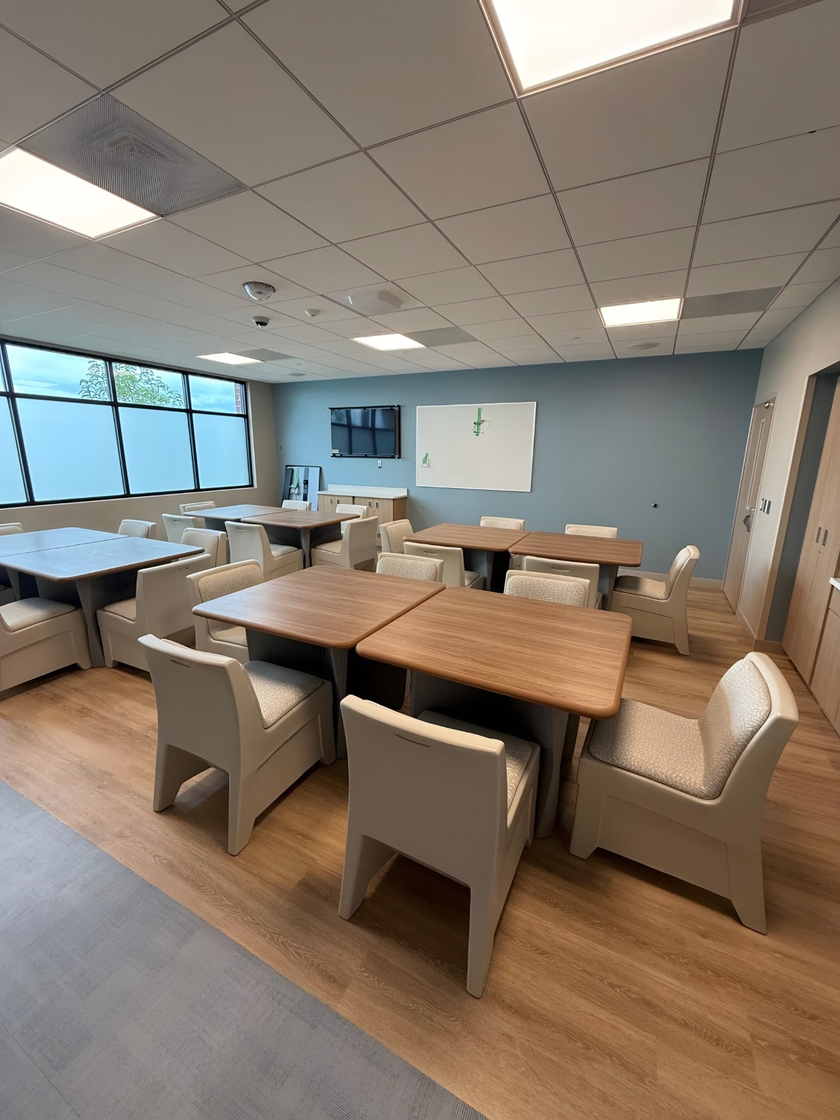 A finished group and dining room in the unit: wood-top tables, tamper-resistant chairs, a blue accent wall and a clerestory window