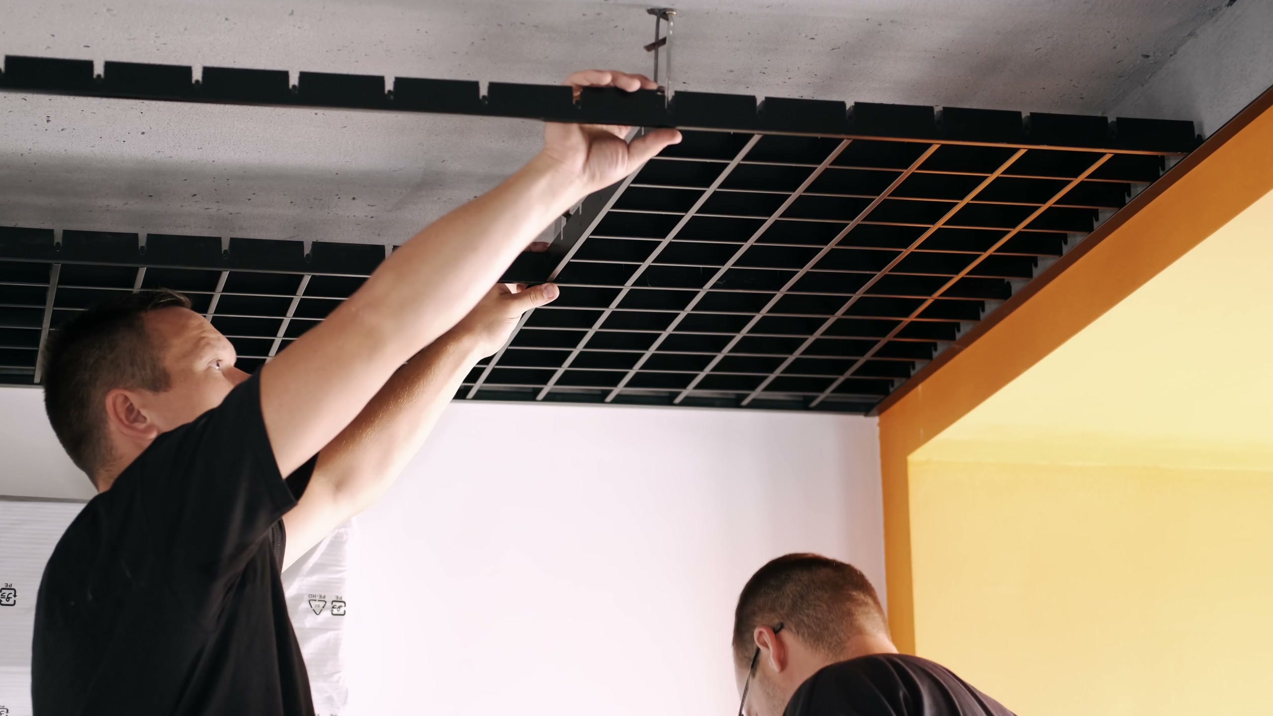 An open-cell grid ceiling going up: a worker in a black T-shirt lifting a section of black grid into place against the slab, a finished run of it overhead and an orange soffit beyond