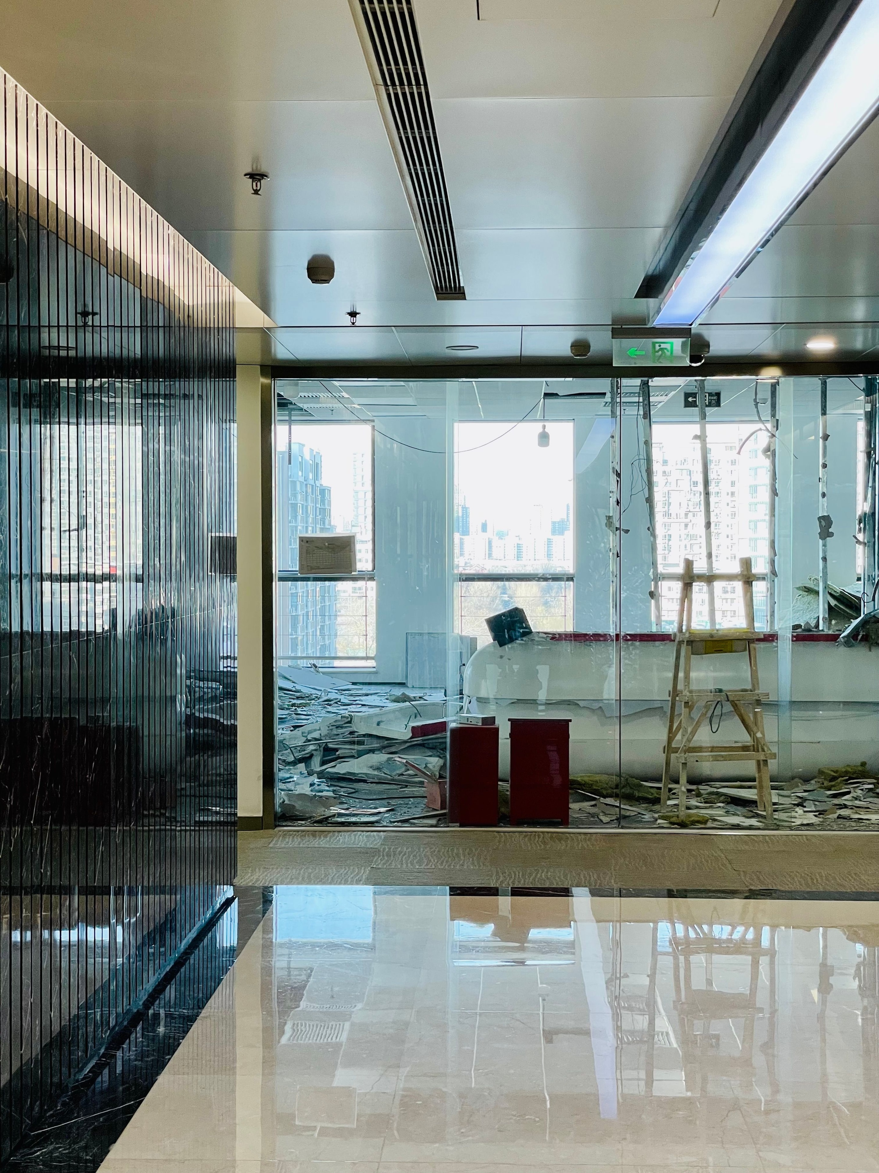 An office floor mid-renovation: glazed partitions along a window wall with a city beyond, ladders and material on the floor, services open overhead