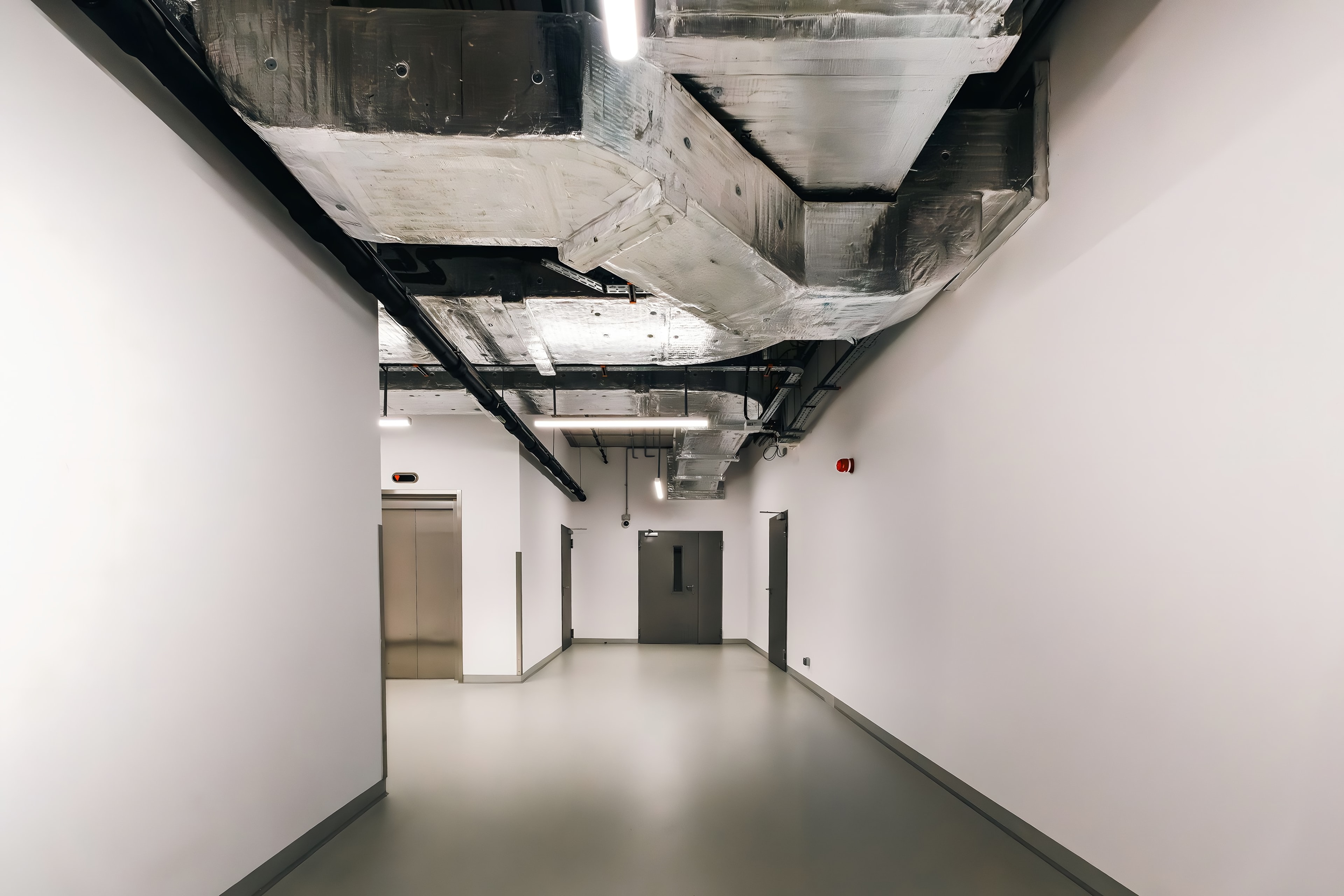 A commercial corridor with its services still open overhead: ducts and conduit above a finished floor, doors and a lift at the far end