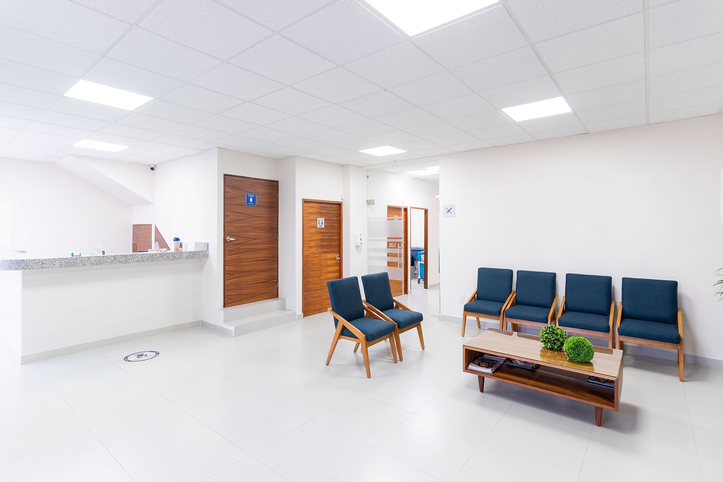 Reception and waiting area in a finished care facility: solid-surface counter, wood doors, seating and a low table under a lit acoustic ceiling