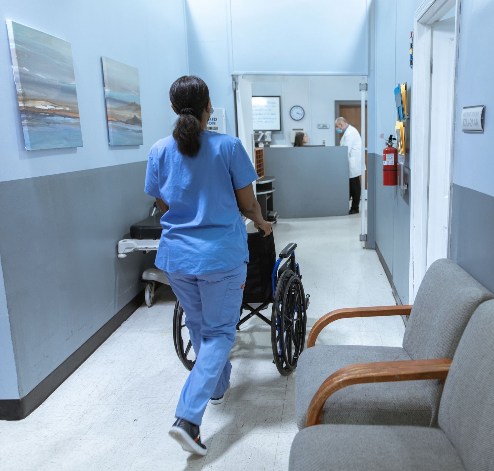 A clinic hallway in service: a nurse in scrubs wheeling a chair past the waiting seats toward the front desk