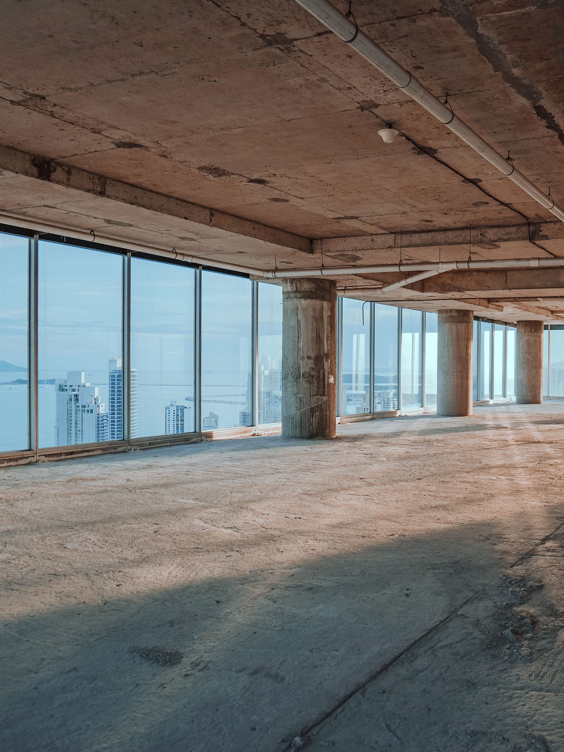 An empty upper floor plate before fit-out: a concrete soffit with conduit run across it, a full-height window wall along two sides with the city and the water beyond, and sunlight raking over the bare slab
