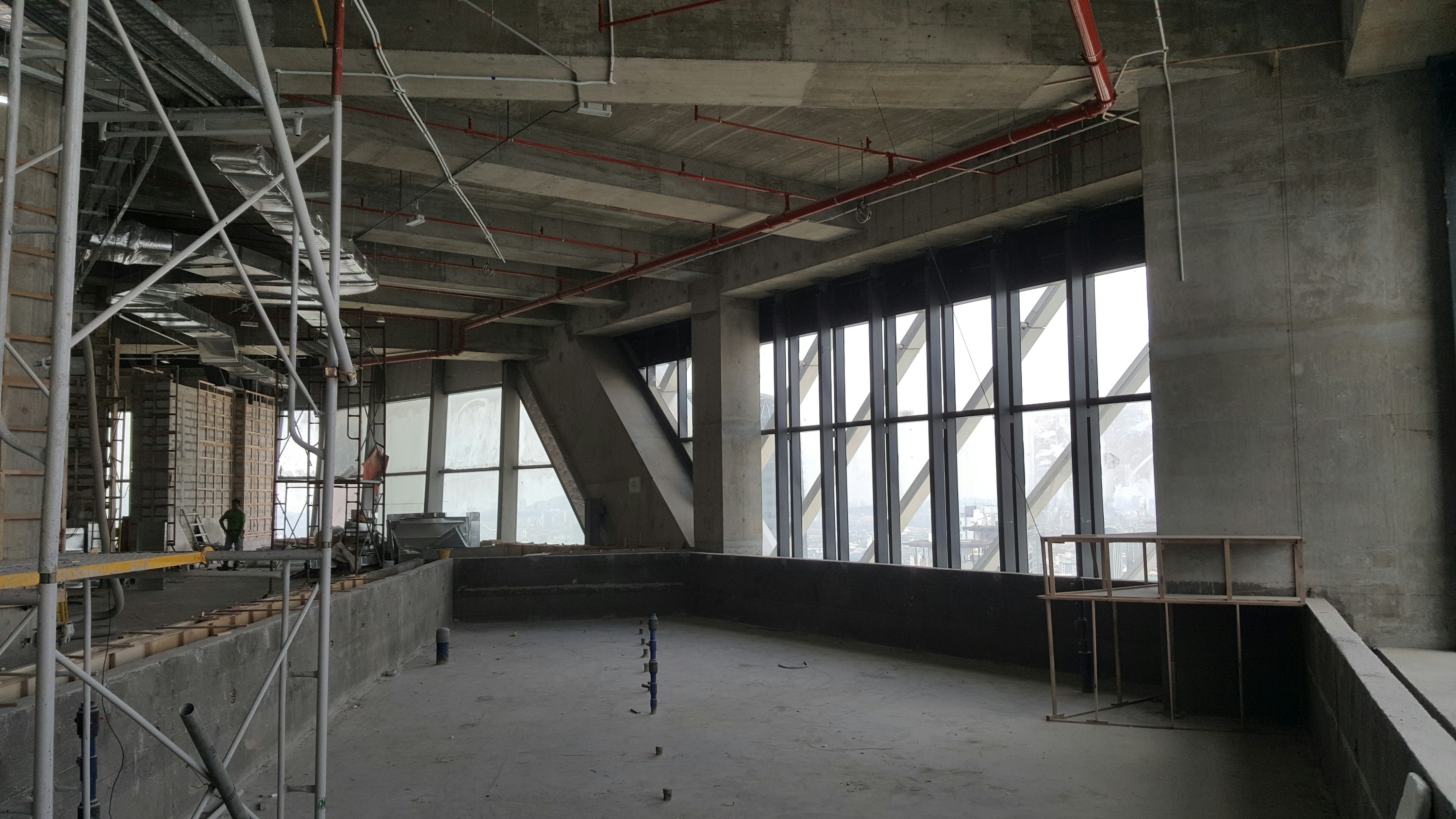 A commercial building interior under construction: a bare slab, concrete columns, ducts and conduit run overhead, scaffolding at the left, and a full-height curtain wall down the right