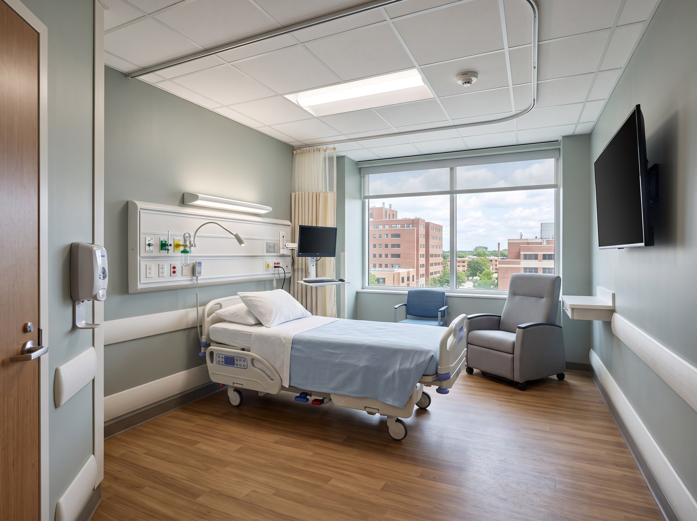 The same patient room returned to service: new bed and headwall, recliner by the window, new floor and ceiling