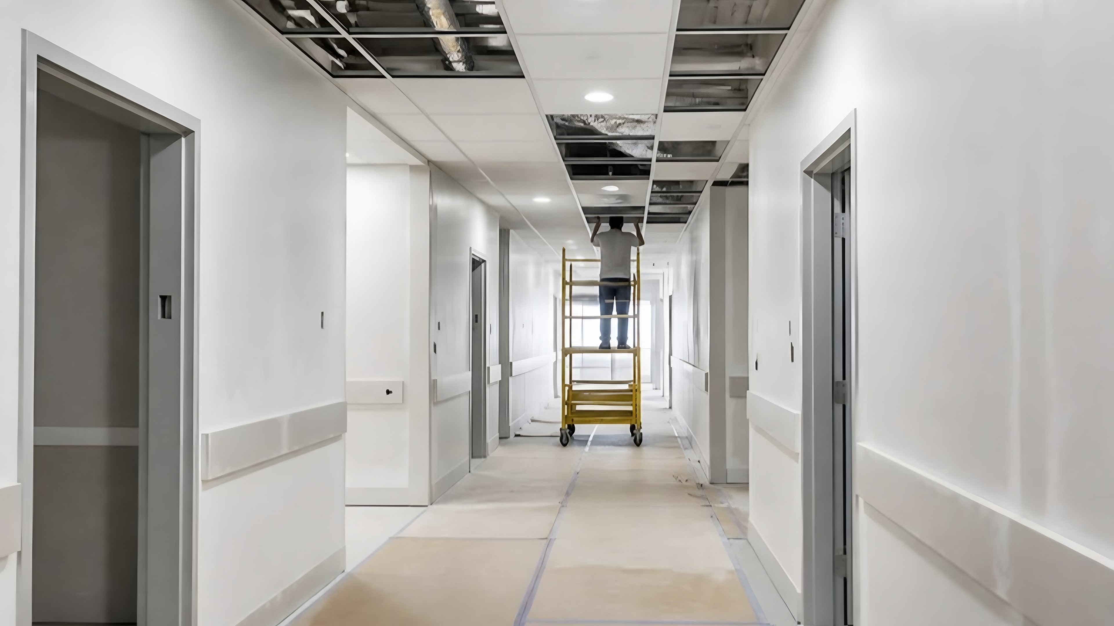 A corridor with finished walls and a suspended ceiling half installed, a worker on a rolling scaffold in the distance