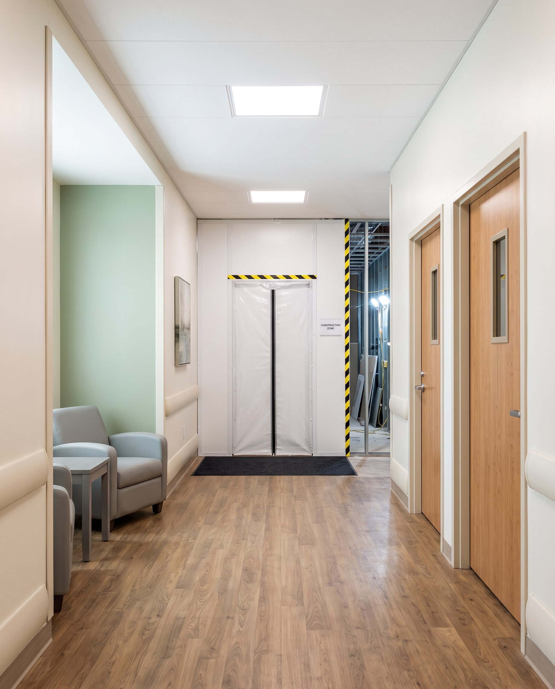 A renovated behavioral health unit corridor that stays open: finished patient room doors and a seating nook, a sealed containment wall with a zippered door closing off the work zone at the far end