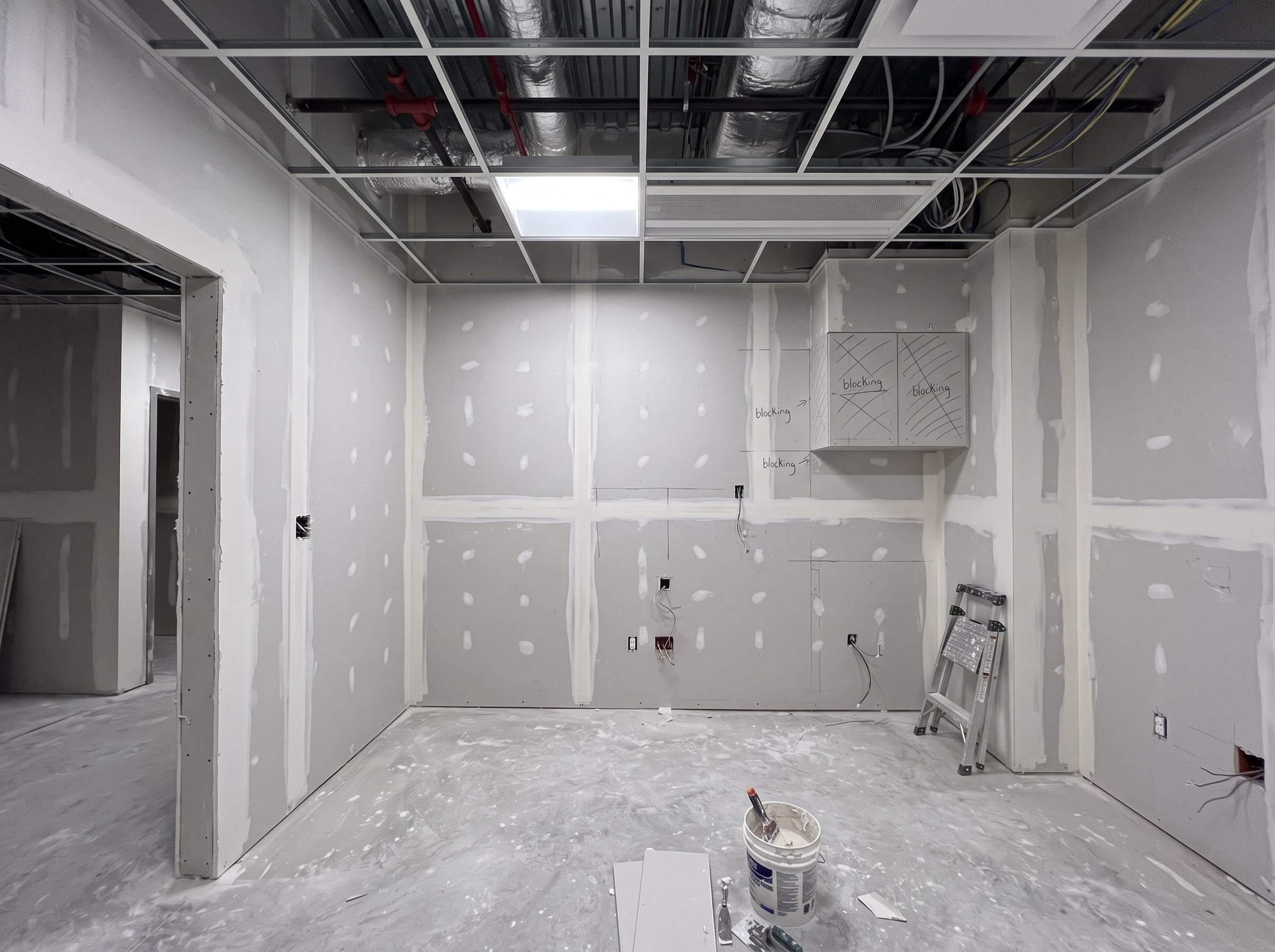 The same exam room after drywall: taped and spotted seams, ceiling grid in, blocking marked where the cabinets will hang