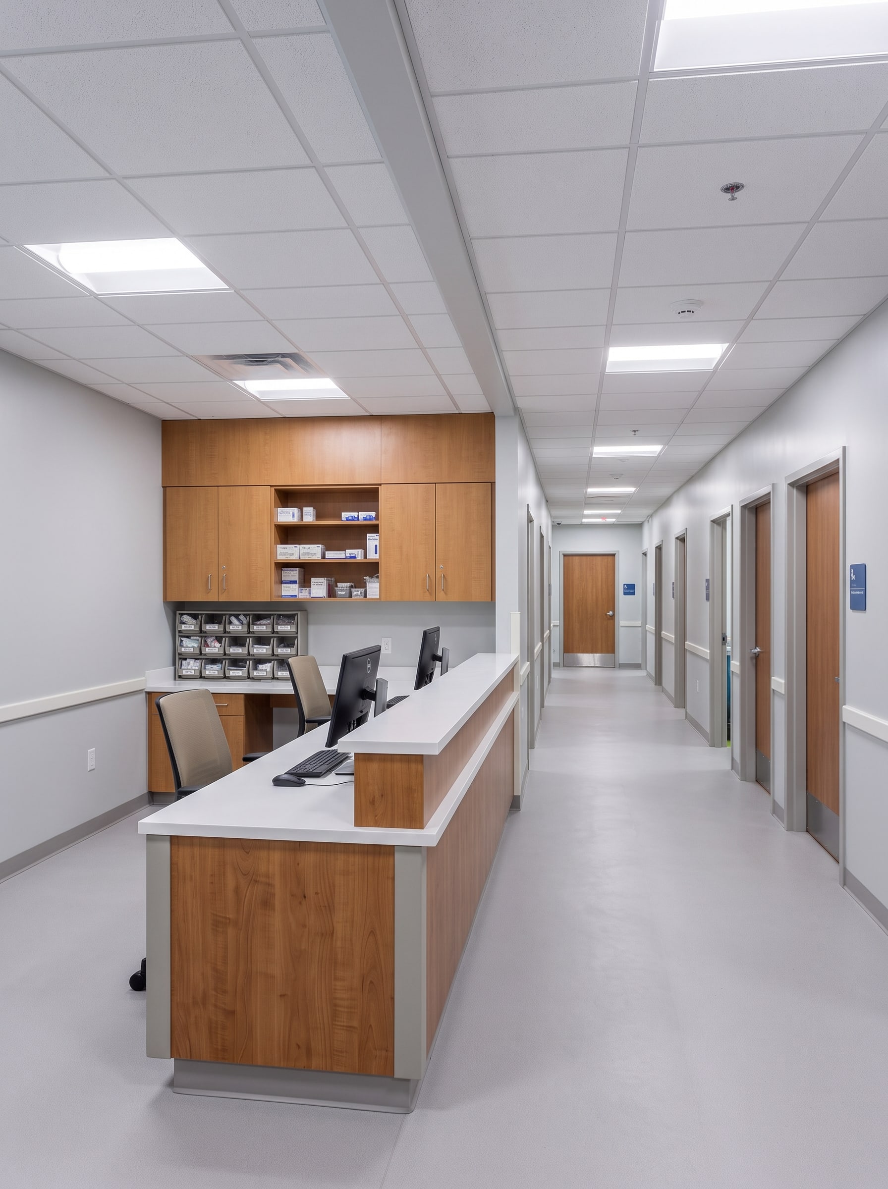 The nurse station on the corridor: two workstations, supply cabinets behind, exam room doors continuing beyond