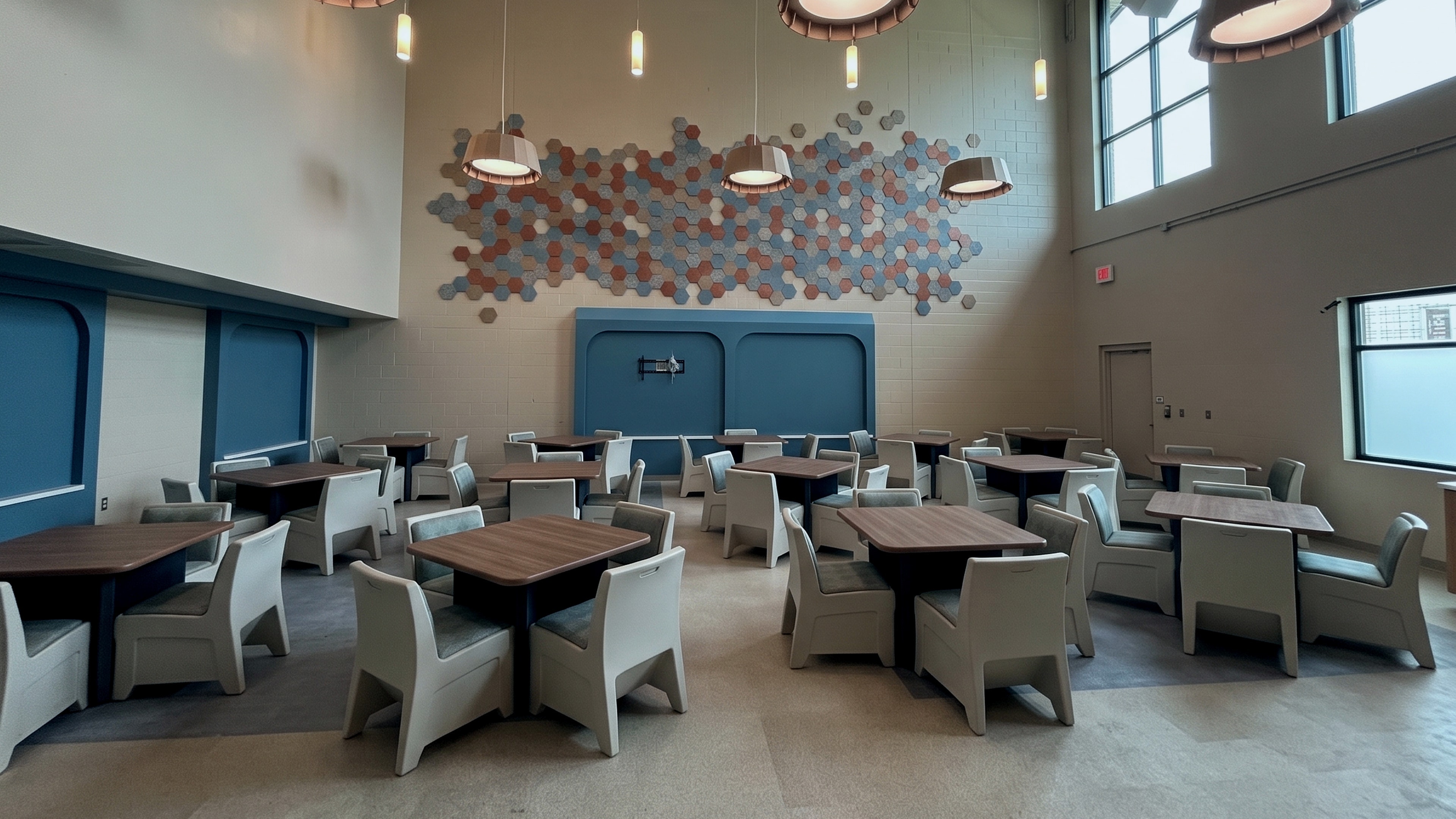 A finished double-height dayroom in a behavioral health facility: tamper-resistant molded chairs at wood-top tables, a blue accent wall, a hexagon acoustic panel feature, drum pendant lights and clerestory windows