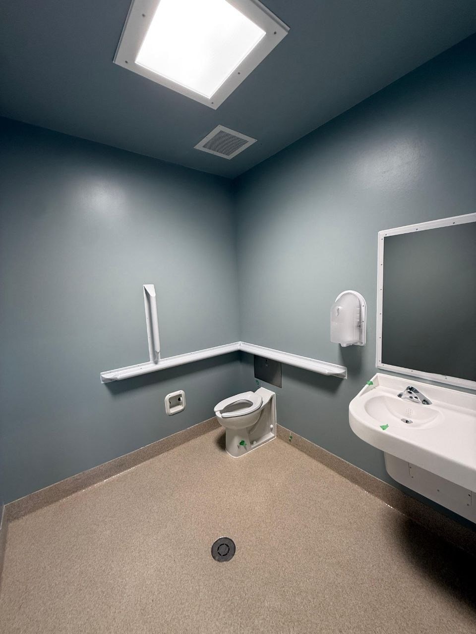 Completed restroom with ligature-resistant fittings: sloped grab bar and shelf, security mirror, seamless resinous floor coved to the wall