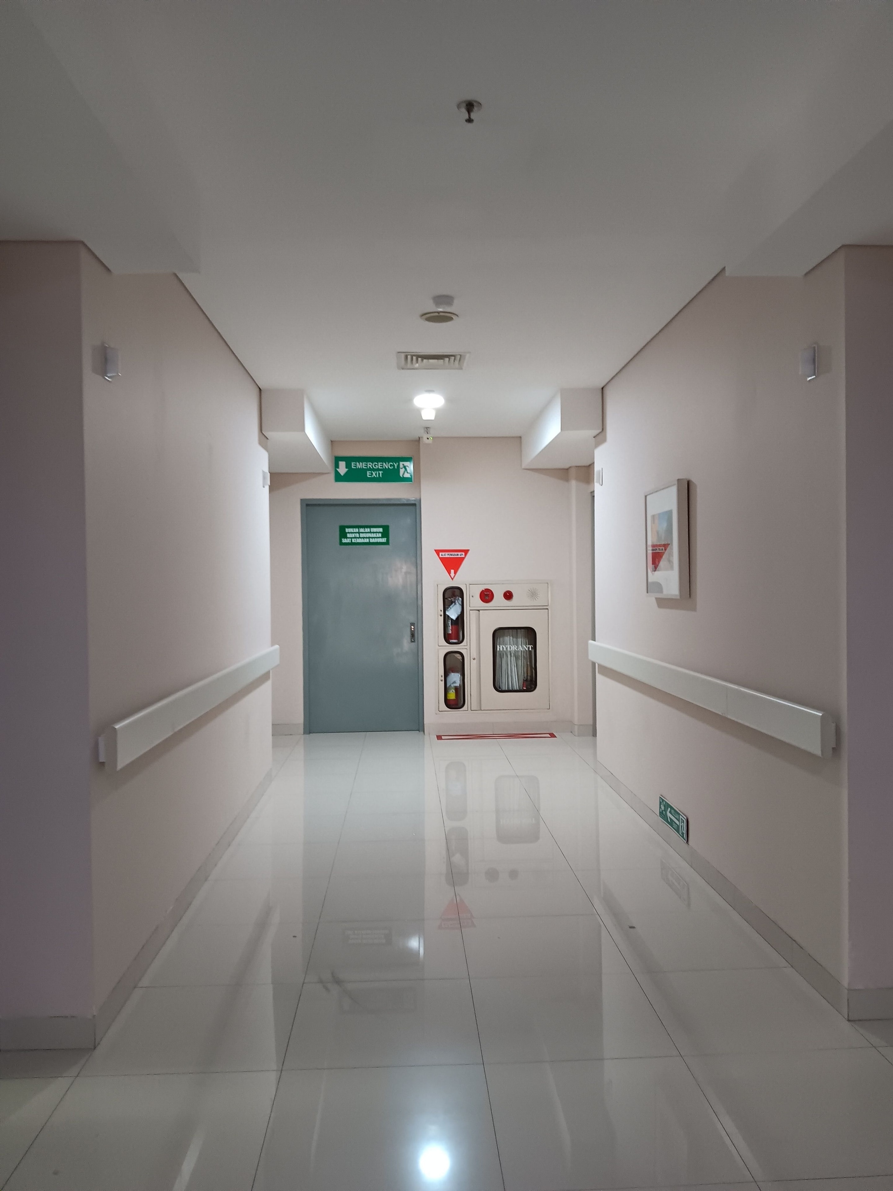 A finished care-facility corridor: handrails along both walls, a glossy resilient floor, an illuminated exit sign and an alarm panel at the far end