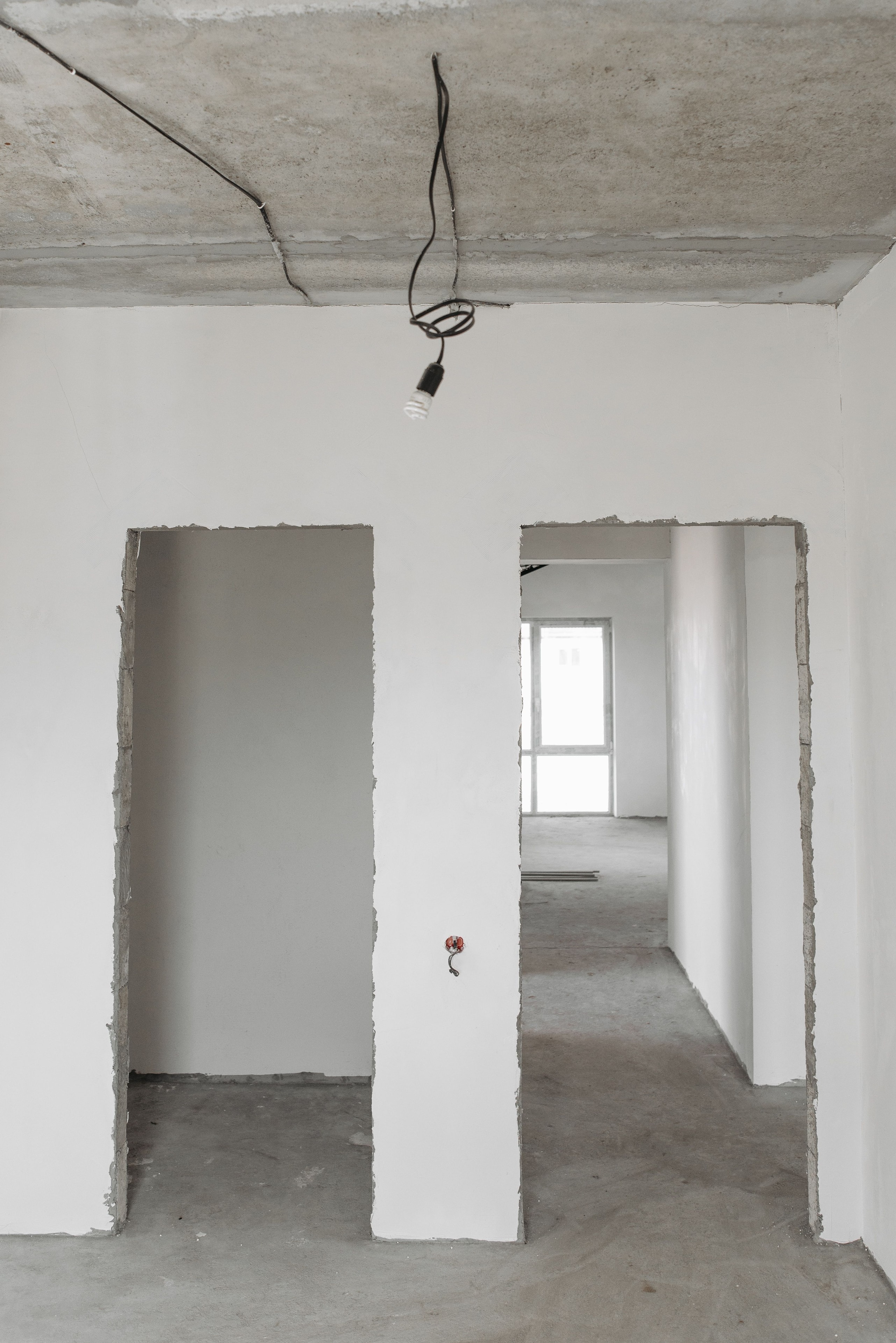 A room in a newly built shell: plastered walls, raw door openings, a bare concrete soffit and a pendant lead hanging from it
