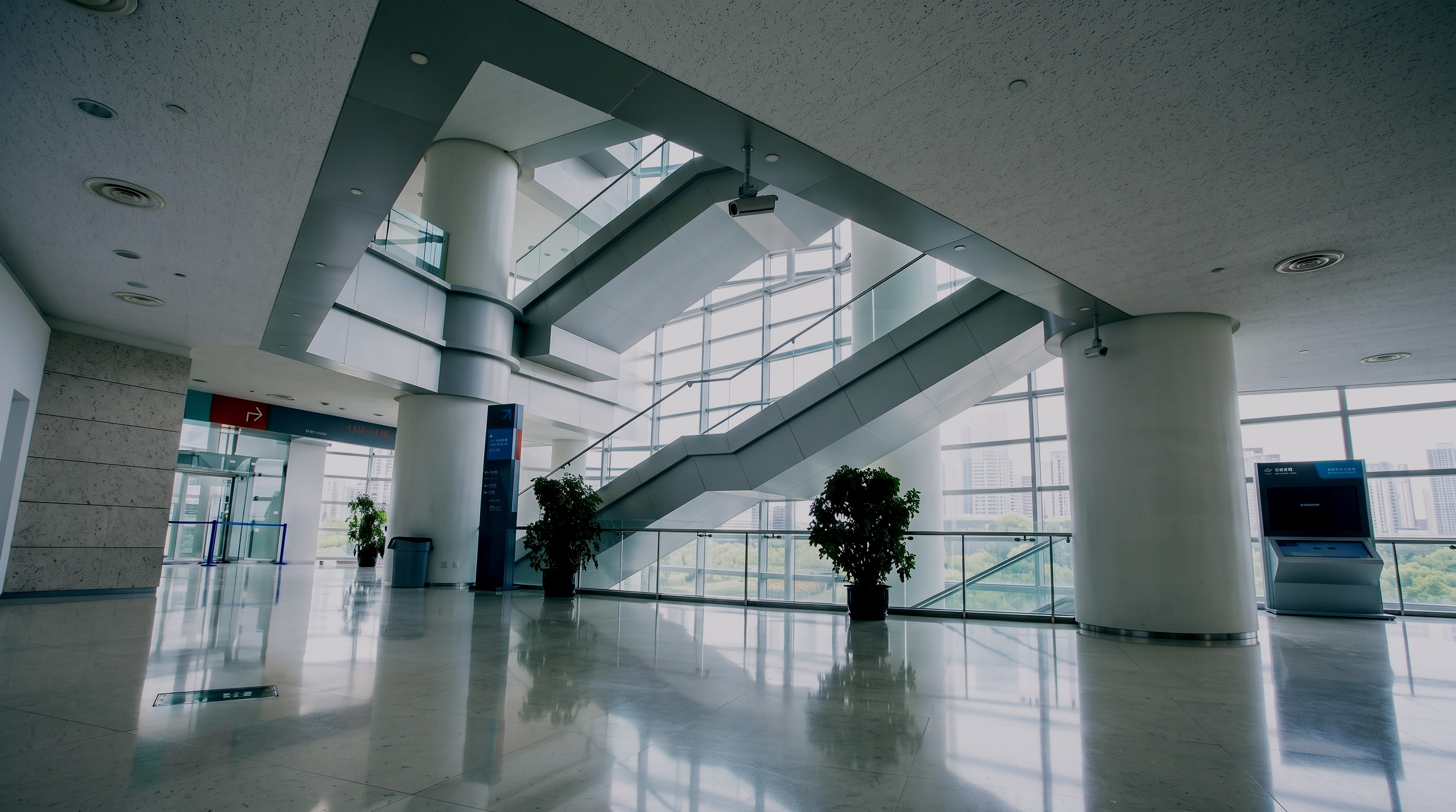 The bright, modern main hall of a healthcare building: crossing stairs under a skylit atrium, round white columns, planters and a polished stone floor before a full-height window wall