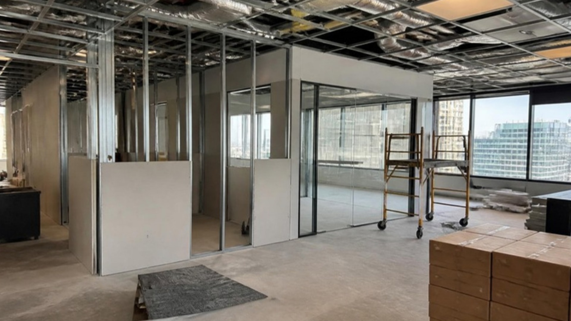 A high-rise office floor mid build-out: metal stud partitions half boarded, a glass-front office going in, a rolling scaffold and carpet-tile boxes by a window wall