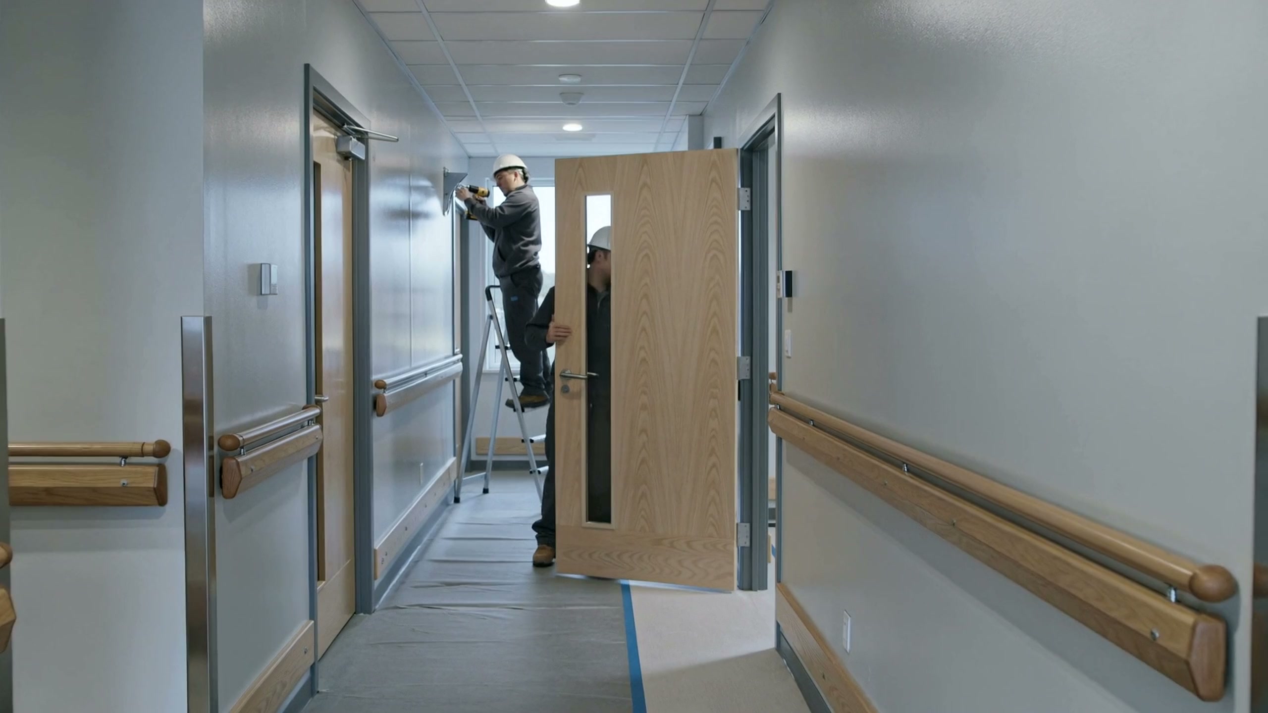 A hospital corridor under renovation: a full-height wood door with a vision panel and a lever handle swinging on the hinges of a steel frame, a worker behind it checking the fit, a second on a stepladder fixing a closer further down, wood handrails along the walls