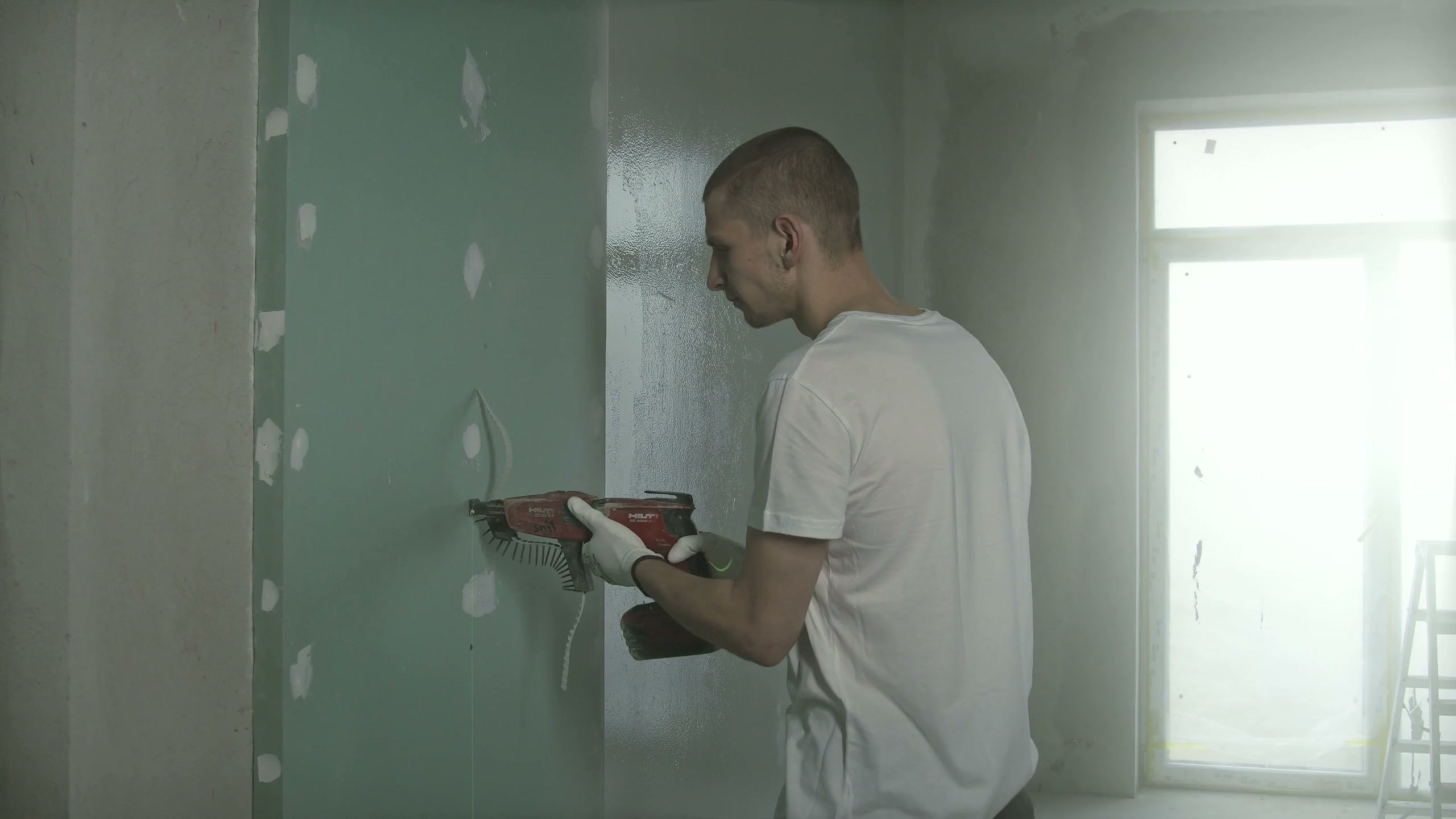 Drywall going up on a metal stud partition: a worker driving screws through a sheet of moisture-resistant board with a collated screw gun, the joints beside him already taped and skimmed