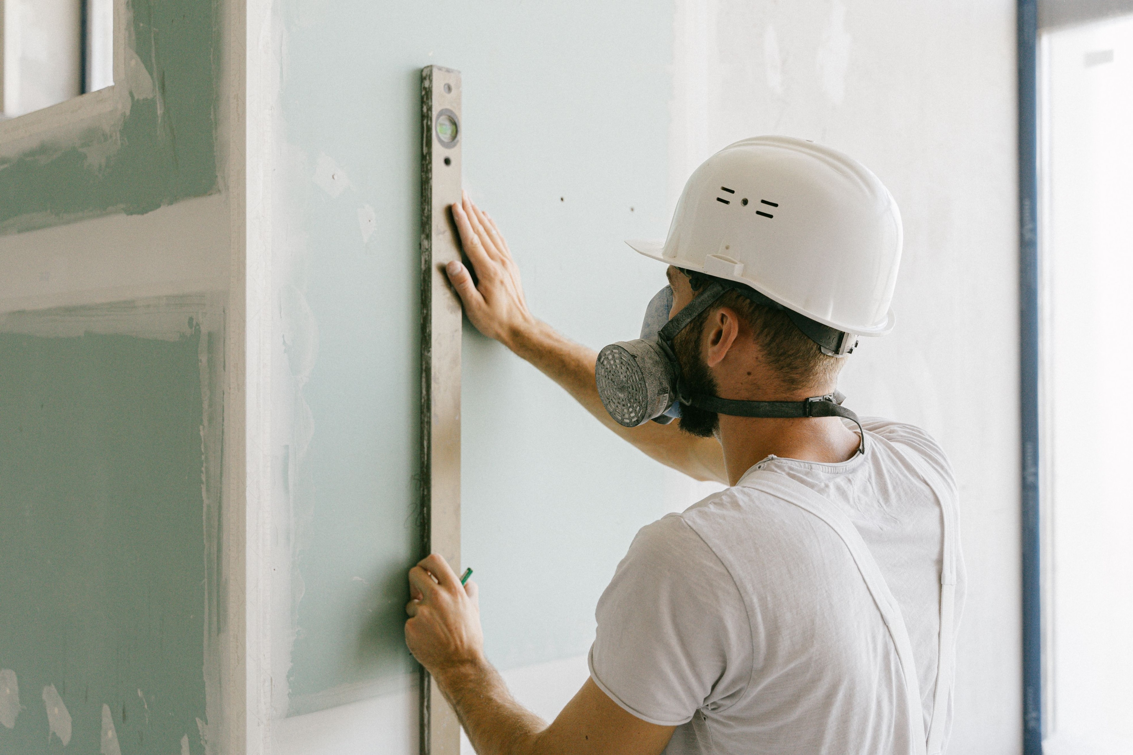 A drywaller in a hard hat and respirator holding a long spirit level against a sheet of green moisture-resistant board, checking that the wall is plumb and flat