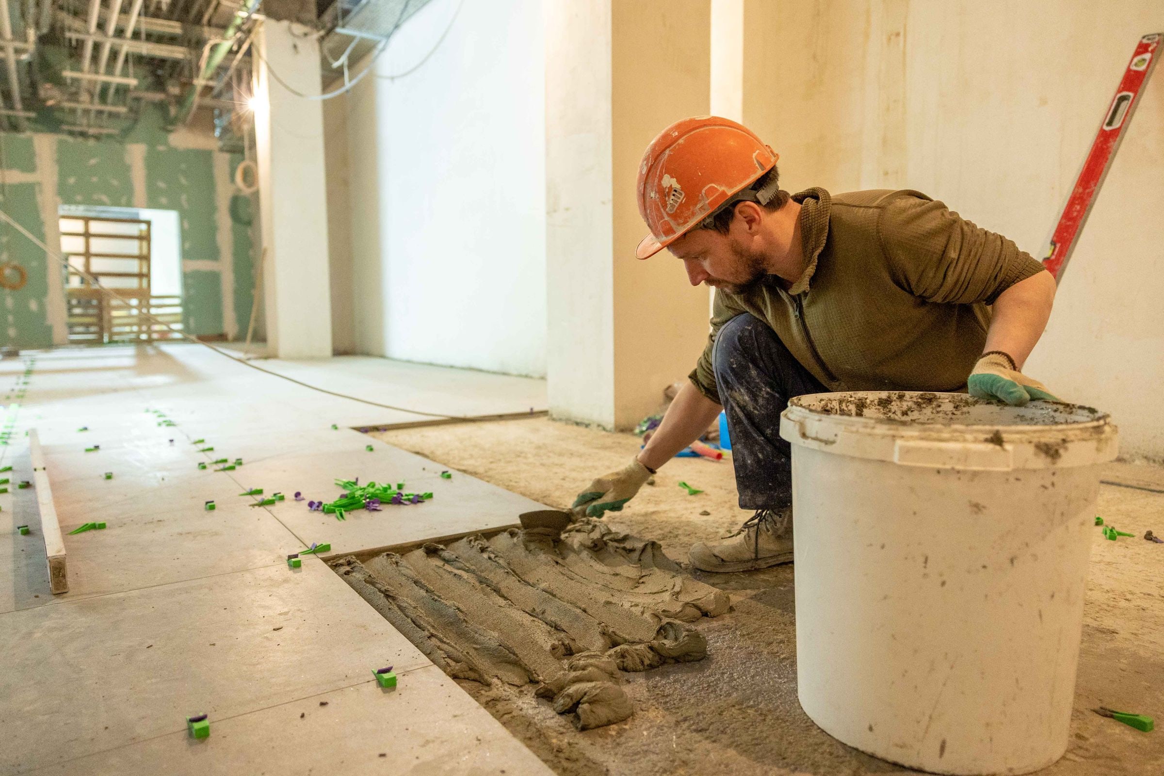 A tile setter in a hard hat on his knees combing thinset across the floor between rows of set tile, a bucket beside him