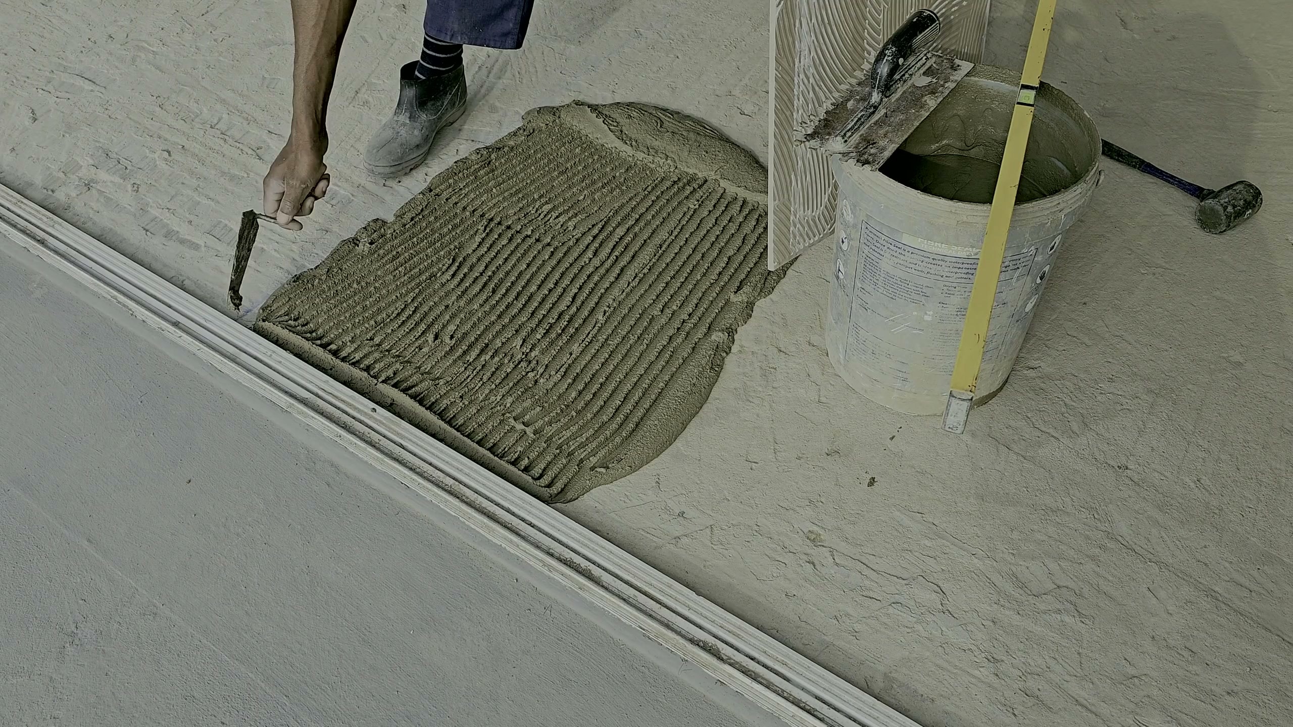 Large-format porcelain tile going down on a commercial floor: a notched bed of adhesive combed out beside a laid run, a bucket of mortar and a margin trowel at the edge