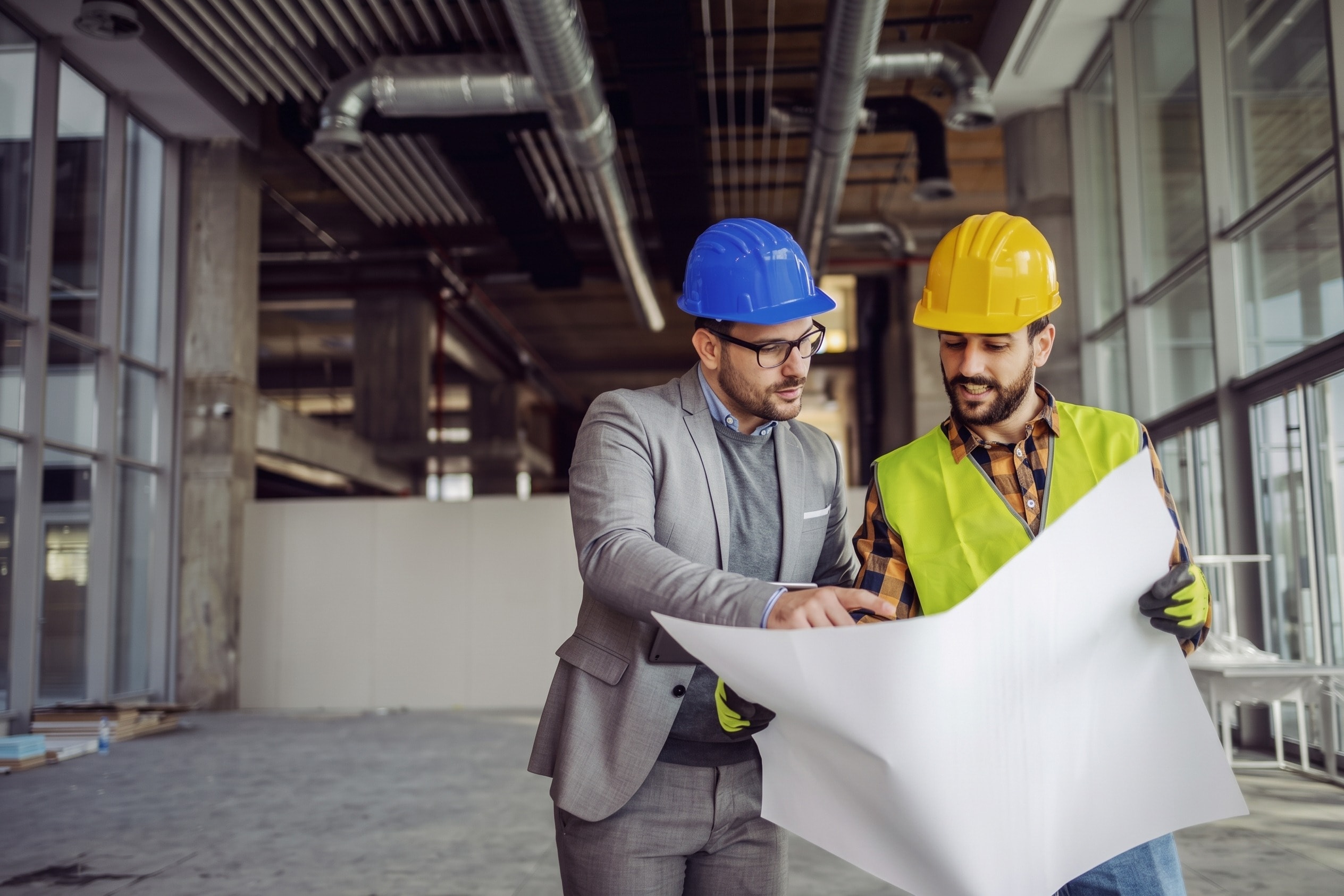 A project manager in a blue hard hat and a foreman in a yellow hard hat and hi-vis vest reviewing a drawing set inside a commercial building under construction, open ceiling services and a glazed facade behind them