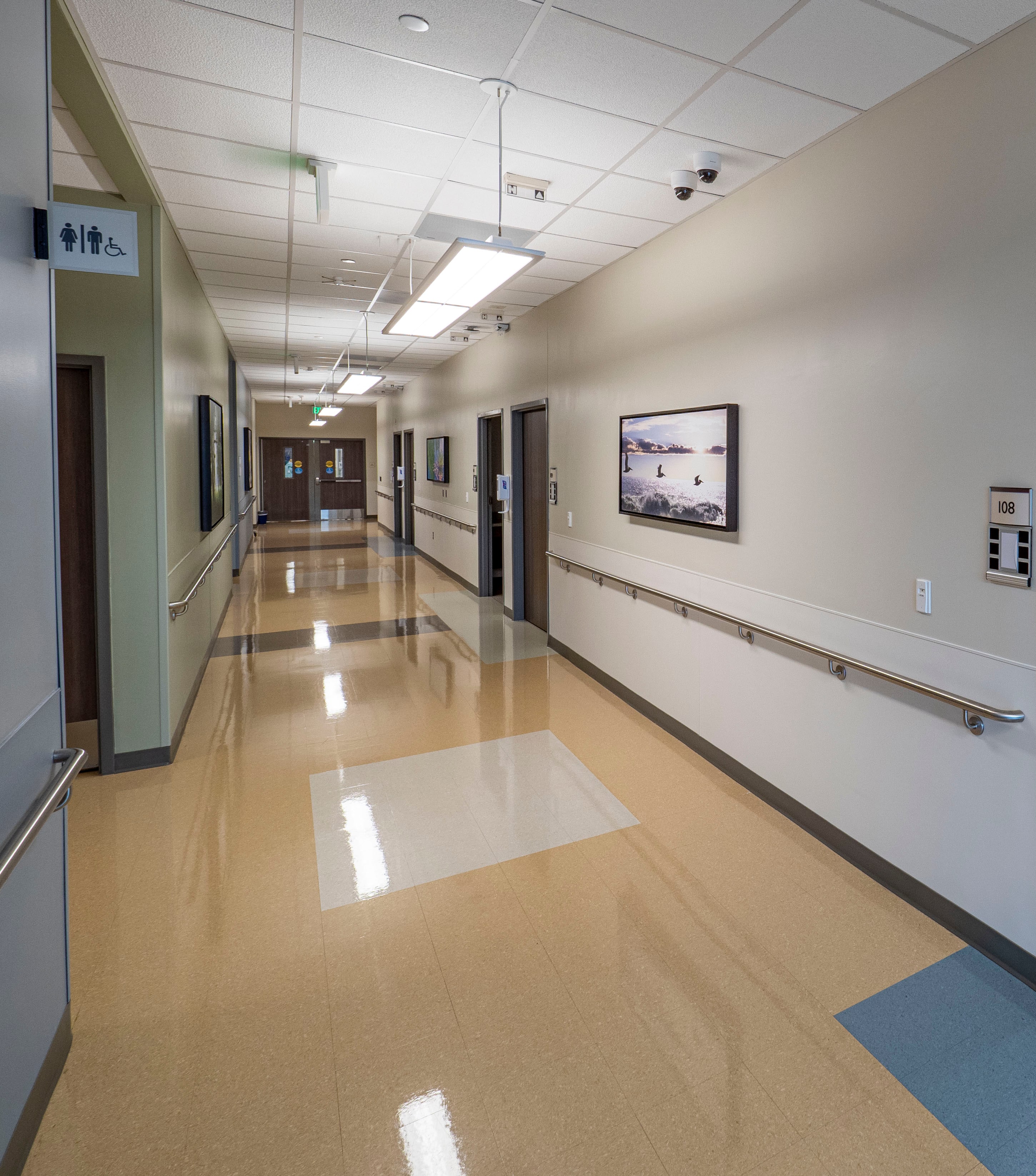 A long, newly finished hospital corridor: polished resilient floor with inset panels, stainless handrails, framed photographs and patient-room doors down the wall, lay-in ceiling with linear lights
