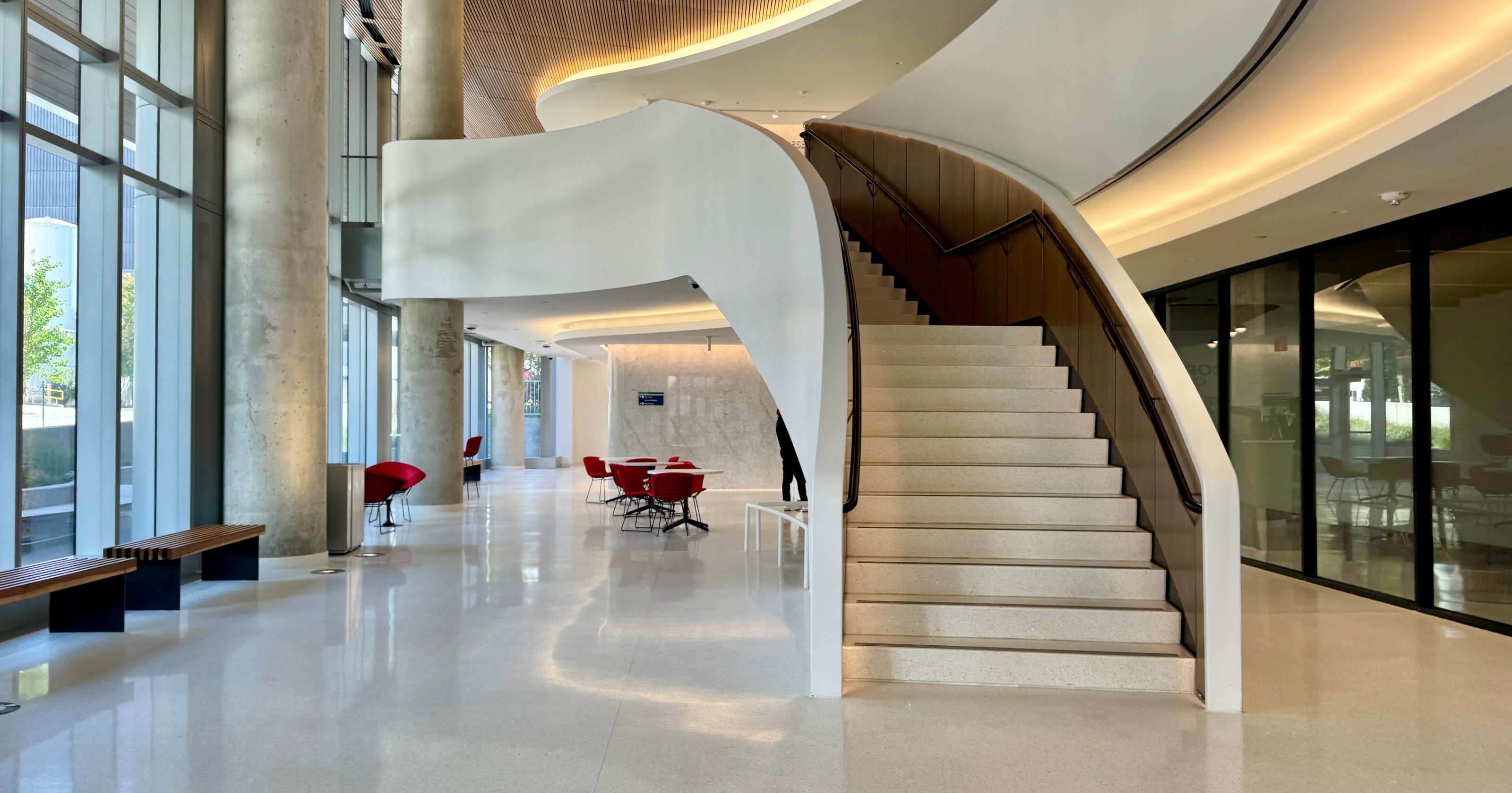 A hospital lobby: a sculptural white spiral staircase with terrazzo treads and a walnut-panelled wall, a slatted wood ceiling with cove lighting, glass walls and a polished terrazzo floor