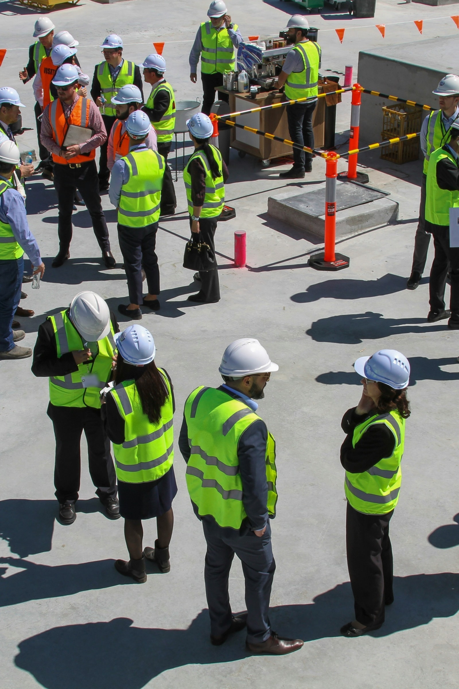Seen from above, a large group in hard hats and hi-vis vests gathered in clusters across a concrete deck for a site briefing, barriers and scaffolding around them