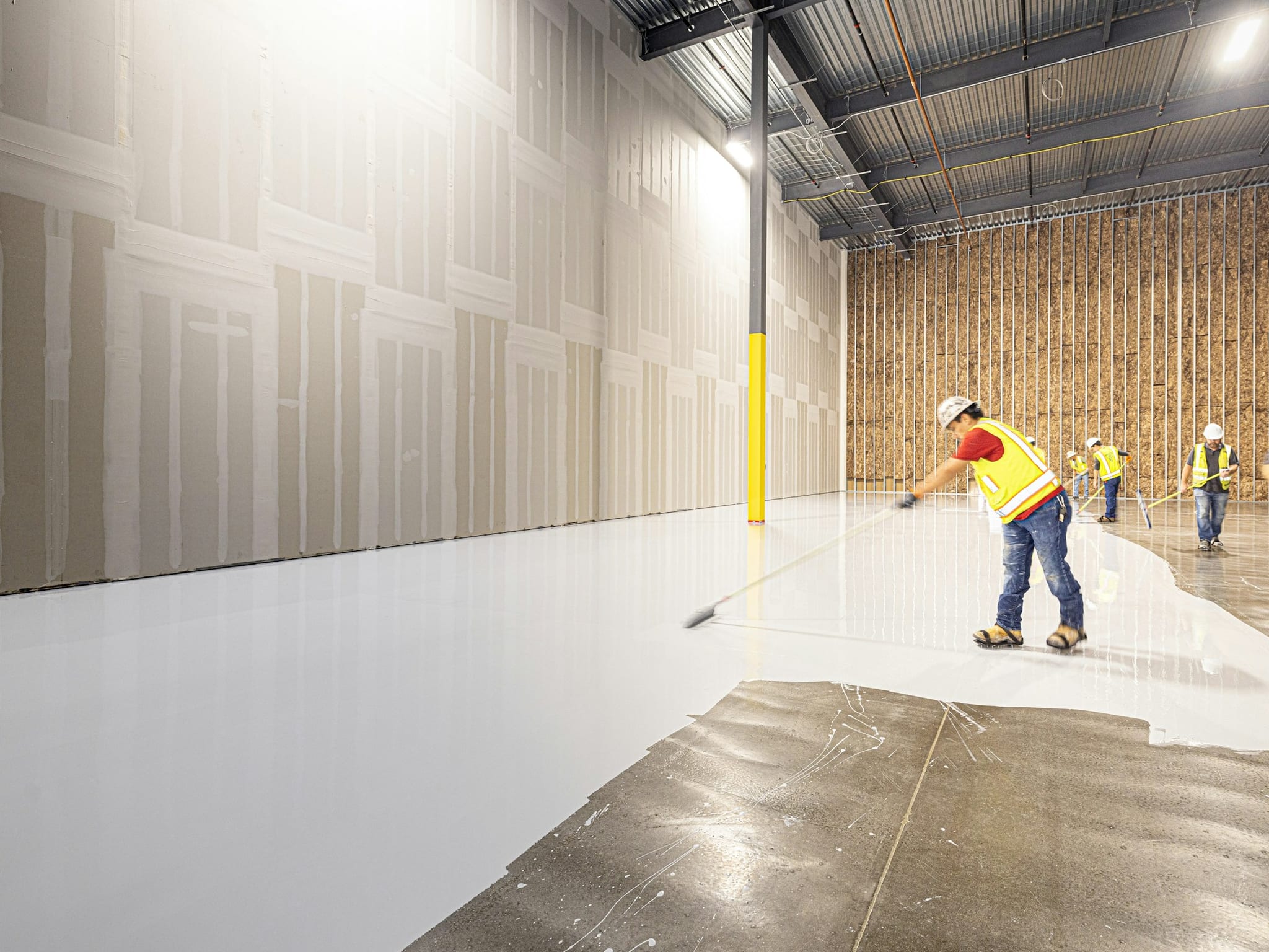 A crew in hi-vis vests and hard hats pulling a resinous floor across a large building, one worker walking the wet edge in the foreground, taped drywall running up to an exposed steel deck behind them