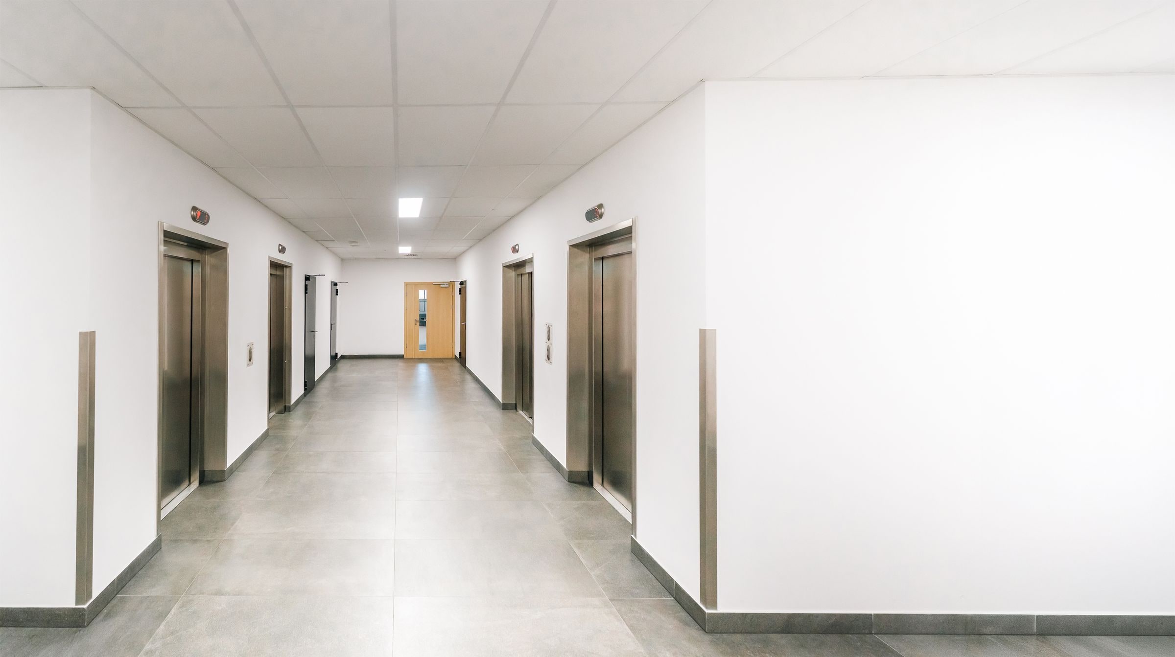 A finished commercial office corridor with stainless corner guards, elevator doors in steel frames and a wood door with a vision panel at the far end