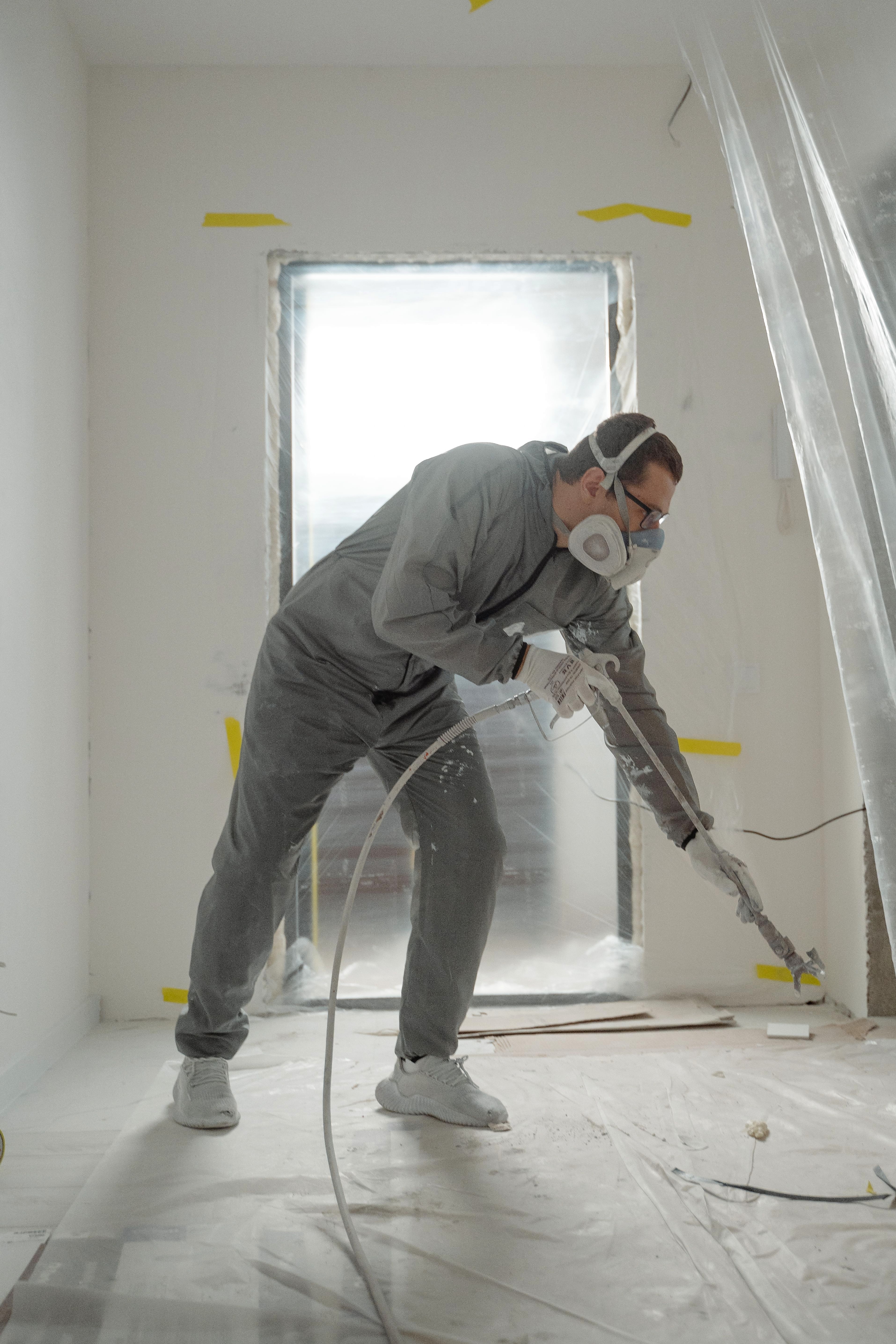A painter in a gray coverall and respirator spraying a wall with an airless gun, the room sealed off with plastic sheeting and the openings masked in yellow tape