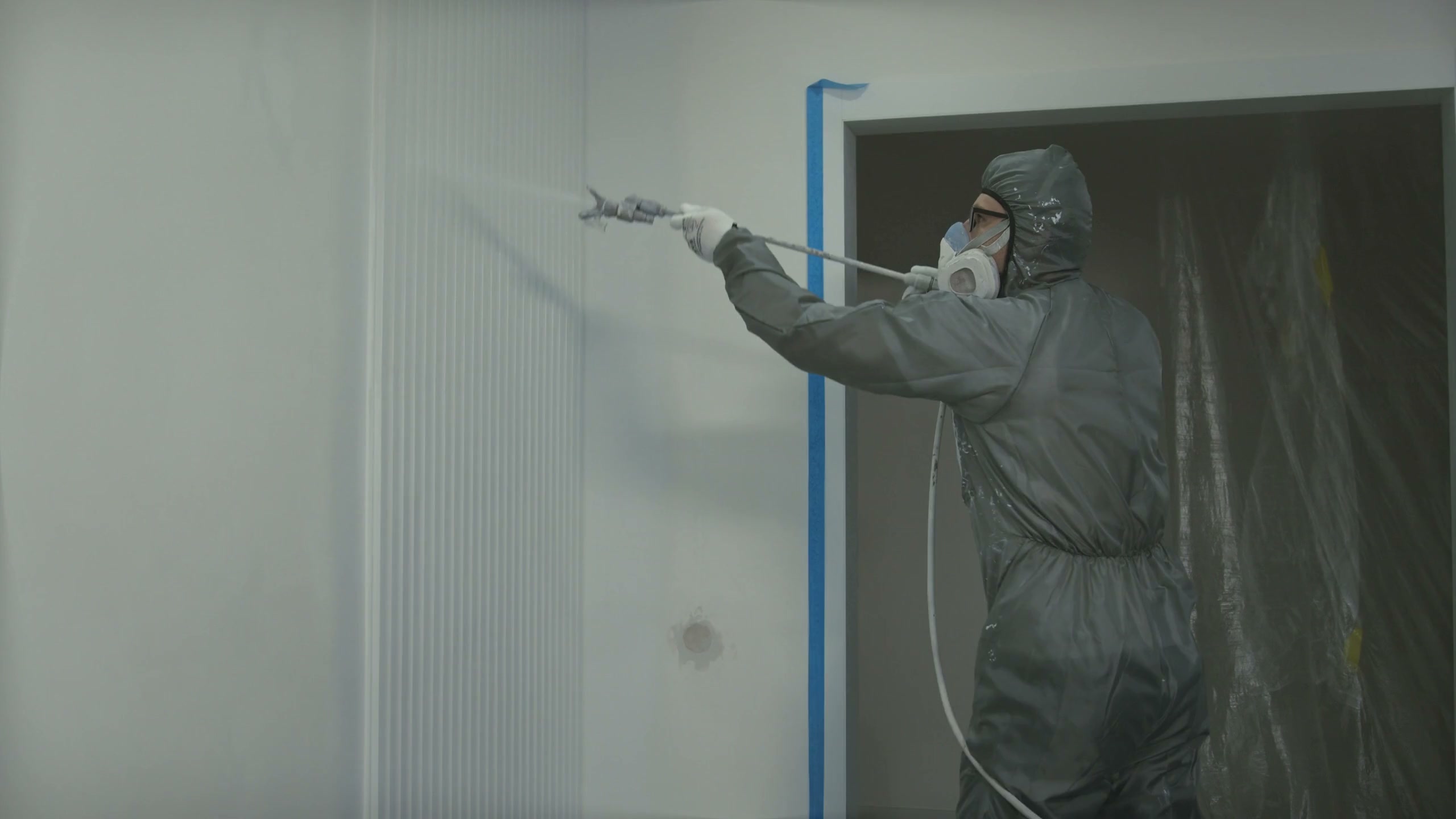 A painter in a coverall and respirator spraying a wall with an airless gun, blue masking tape run down the edge of a door frame beside him