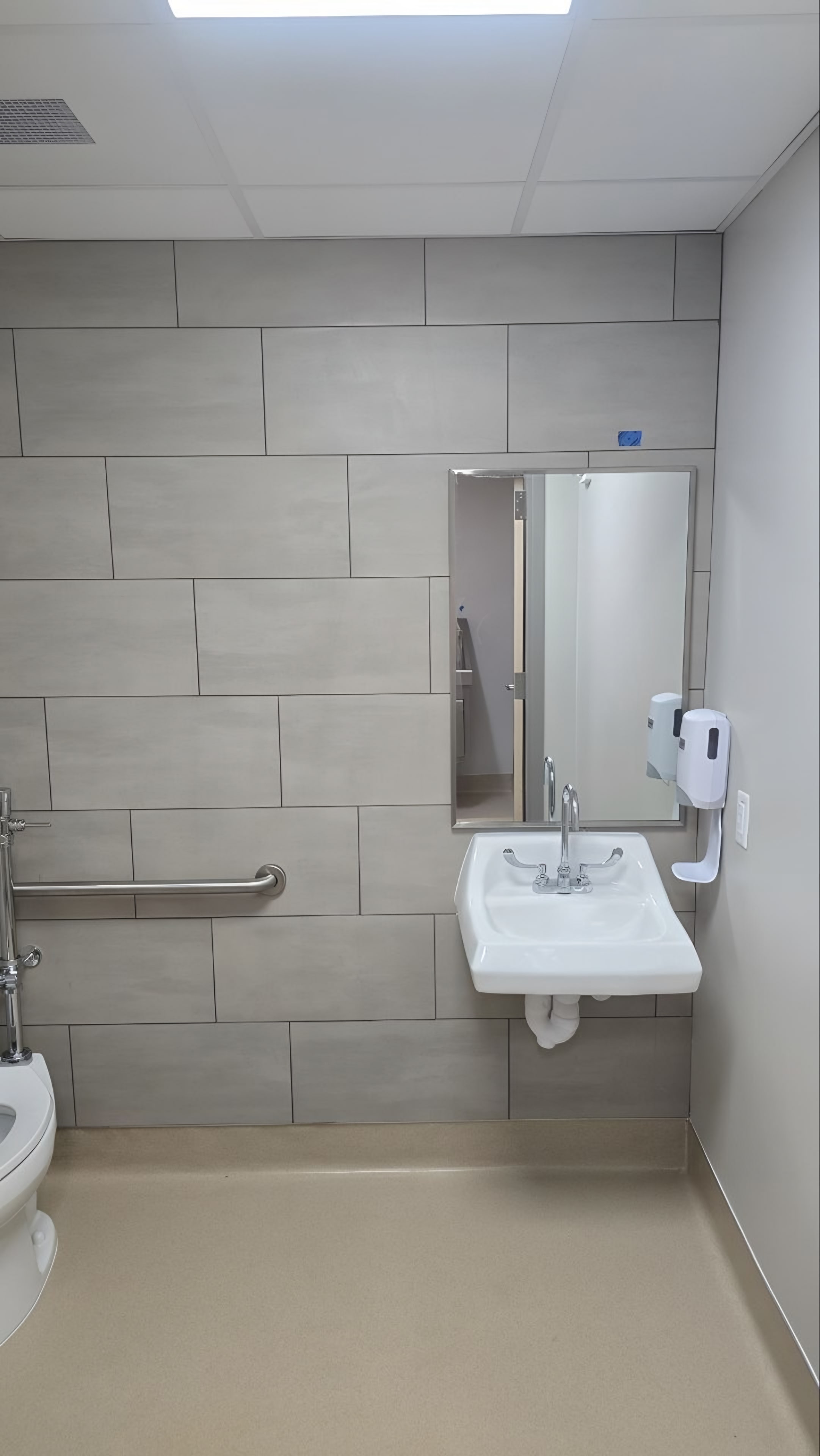 A restroom in large-format grey tile with a wall-hung lavatory and grab bars