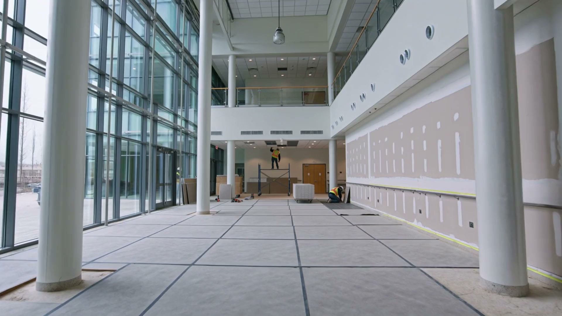 The double-height lobby of a hospital under interior renovation: a glass curtain wall on the left, a mezzanine with a glass railing above, a worker on a rolling scaffold at an open ceiling grid, stacks of drywall and a taped board wall with a handrail on the right