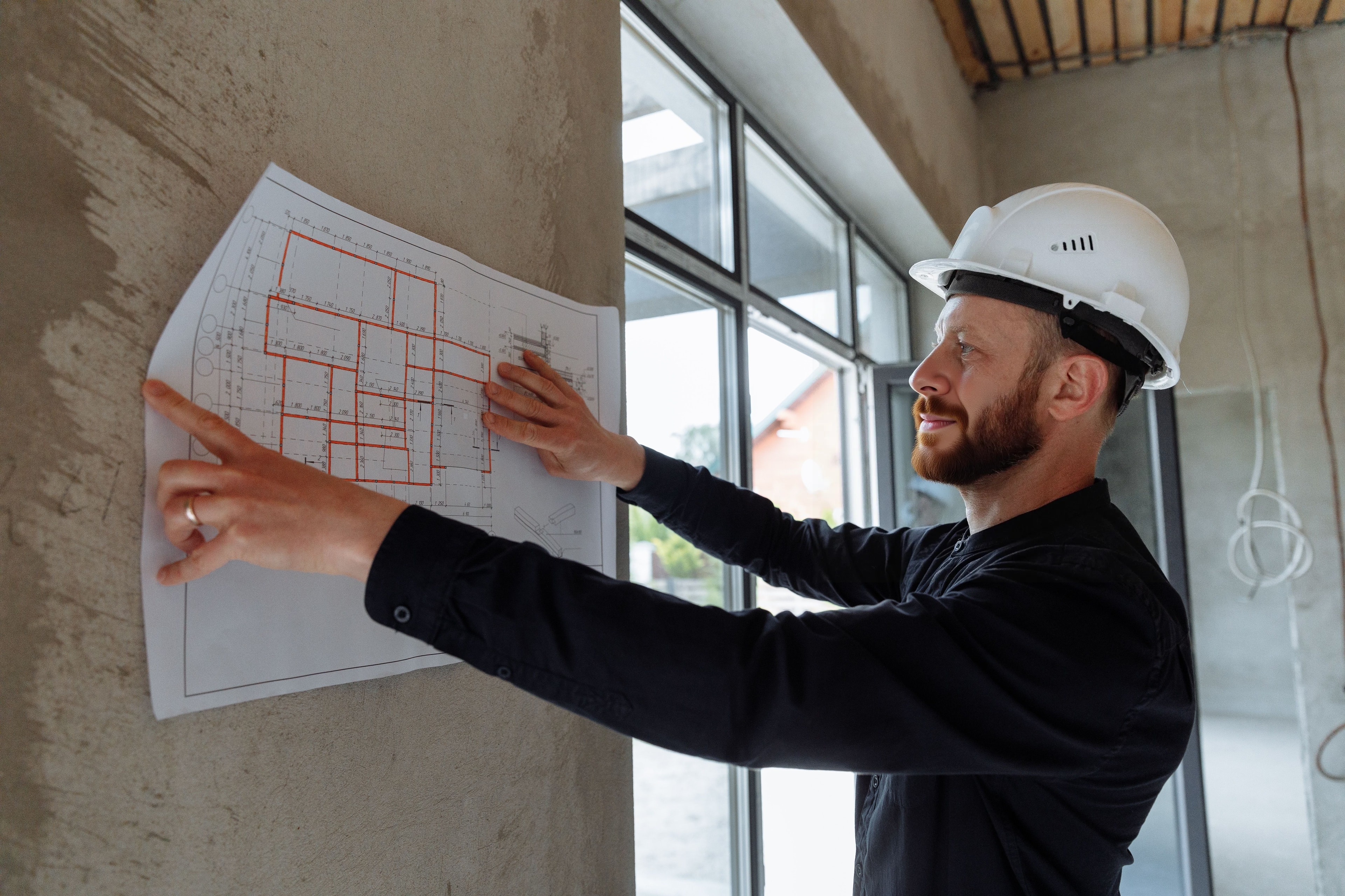 A supervisor in a white hard hat pressing a floor plan flat against a concrete column beside a window wall, one finger on the partition layout marked out in red