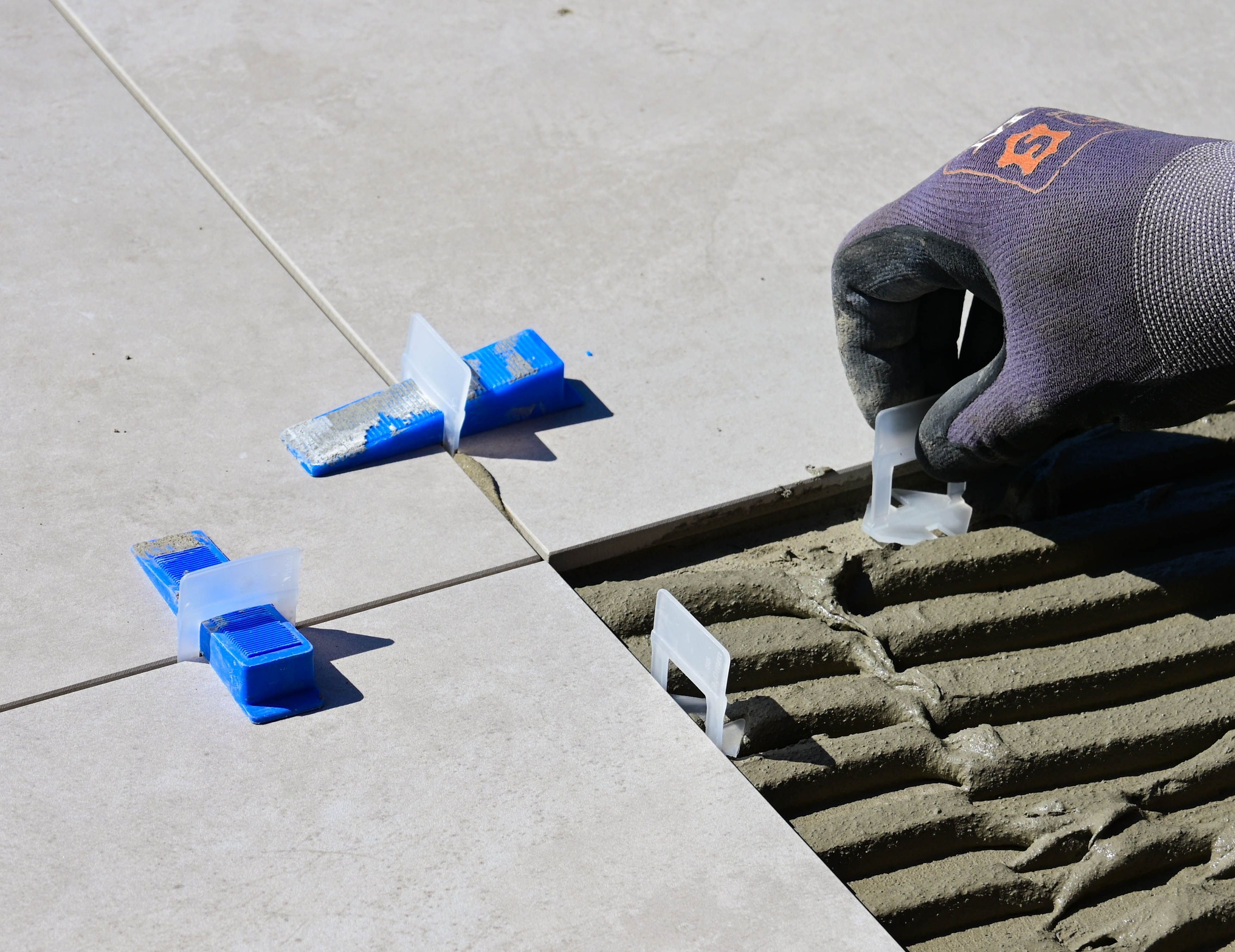A gloved hand setting tile leveling clips between large-format porcelain tiles, the ridged thinset bed for the next tile beside them